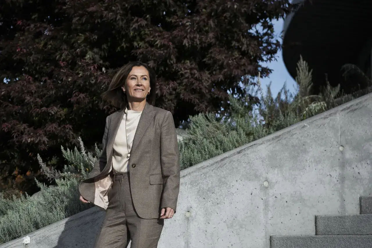 Helena Hedblom i en brun kostym, med en ljusblus och ett pärlhalsband, står utomhus vid en betongtrappa med grönska och träd bakom sig.