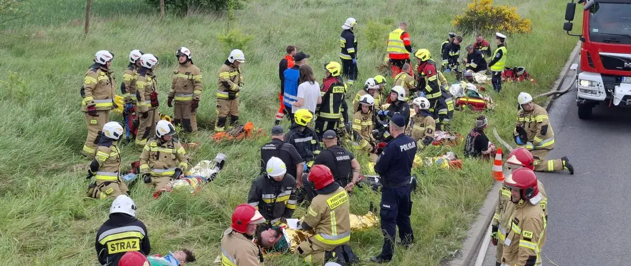 Strazak w akcji, ratuje poszkodowanych po wypadku. Wokół niego inni strażacy i policja.