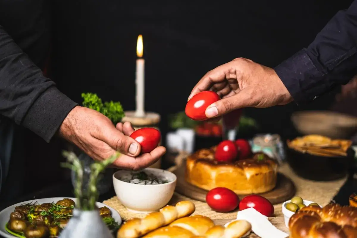 Hands exchange red Easter eggs, a tradition celebrated when is Easter in Europe. A candle flickers behind a festive spread.