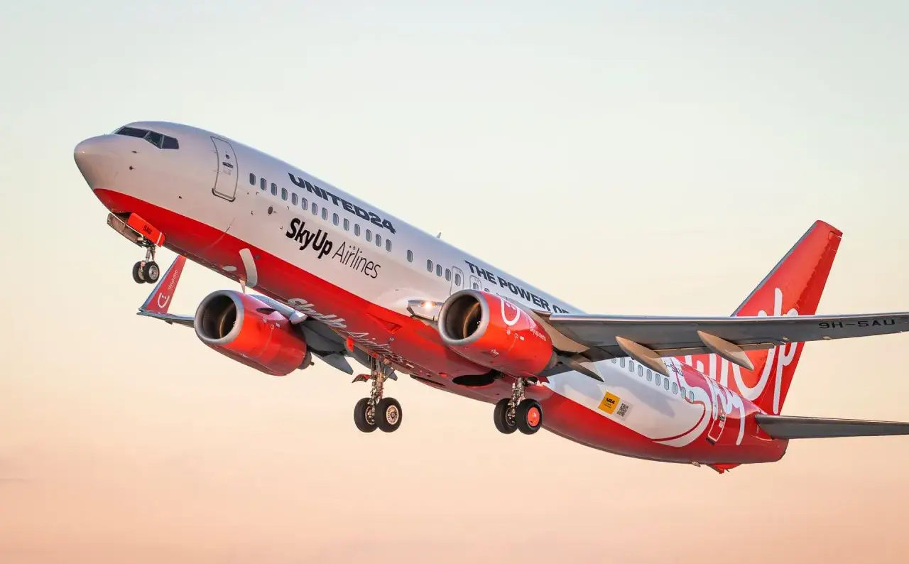 Samolot SkyUp Airlines wznosi się w powietrze. Czerwono-białe barwy maszyny i logo linii lotniczych są dobrze widoczne na tle jasnego nieba.