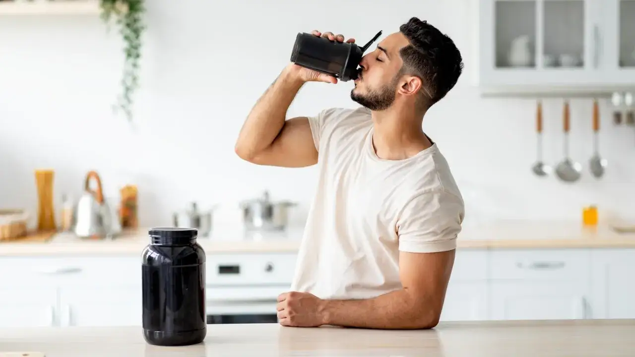 Mann trinkt proteinshake vor dem schlafen aus einem schwarzen Shaker. Daneben steht ein gro&szlig;er Beh&auml;lter mit Pulver.