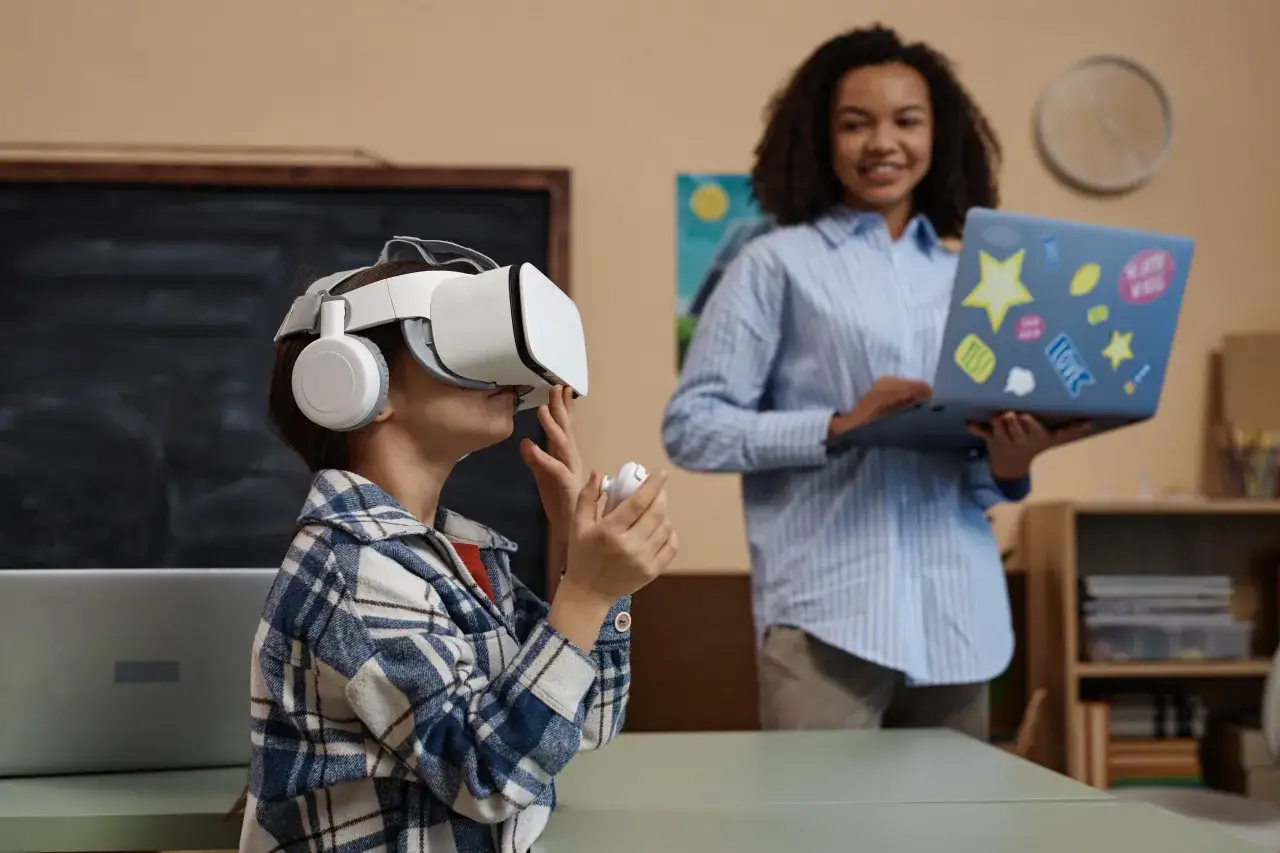 Dziecko w goglach VR odkrywa wirtualny świat, podczas gdy nauczycielka z laptopem obserwuje. To pokazuje, co to jest VR &ndash; technologia immersji.