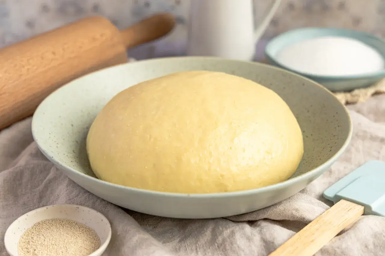 Hefeteig in Schüssel, daneben Trockenhefe. Frage: wieviel trockenhefe statt frischer hefe? Teig, Zucker, Nudelholz, Spatel.