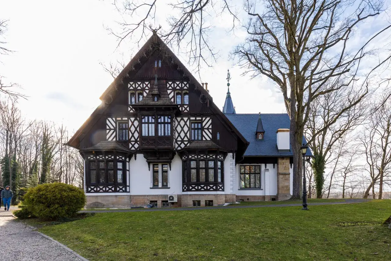 Pałac myśliwski w Promnicach, z charakterystyczną architekturą szachulcową i porośniętym mchem dachem, otoczony drzewami.