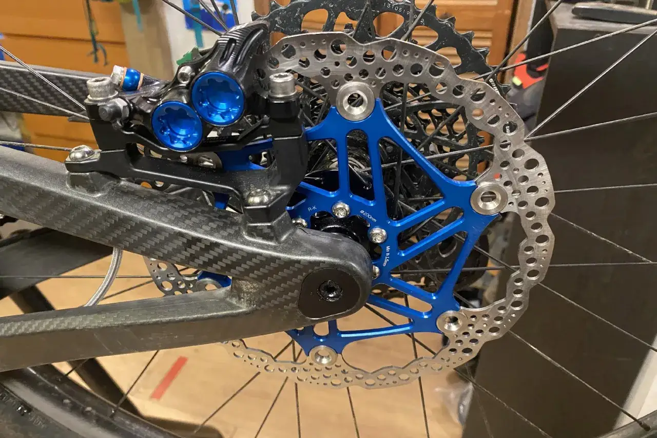 Close-up of a bicycle's rear brake system, showing a carbon fiber frame, blue brake caliper, and disc rotor. A silver hydraulic hose is visible, ready for cutting.