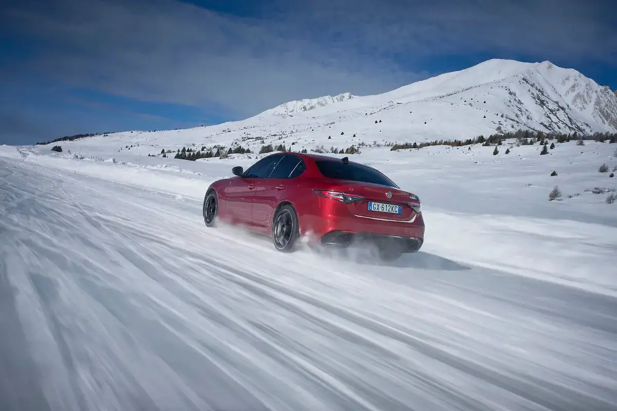 Czerwone Alfa Romeo Giulia Q4 z napędem na cztery koła śmiga po ośnieżonej drodze, wzbijając śnieg. W tle góry i błękitne niebo.