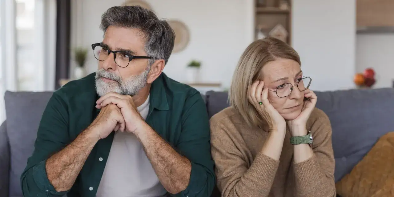 Ein &auml;lteres Paar sitzt auf einem Sofa, beide mit nachdenklichem, traurigem Blick. Ihre K&ouml;rpersprache deutet auf eine lieblose Ehe im Alter hin, voller unausgesprochener Worte und Distanz.