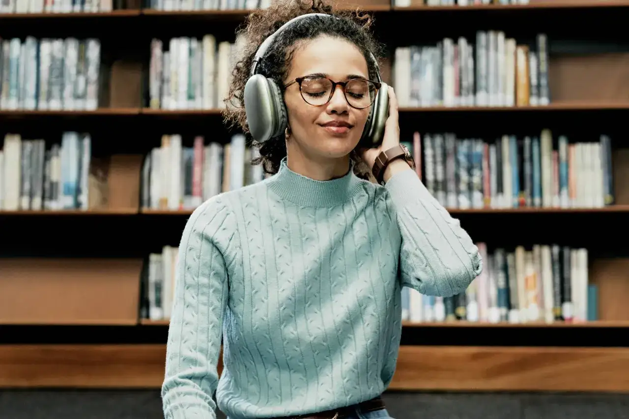 Kobieta w okularach i słuchawkach na tle regał&oacute;w z książkami, słucha najlepszych audiobook&oacute;w.