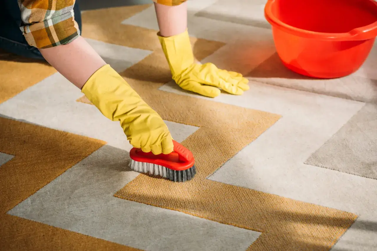 Manos con guantes amarillos frotan una alfombra con un cepillo rojo. Un cubo naranja está cerca.
