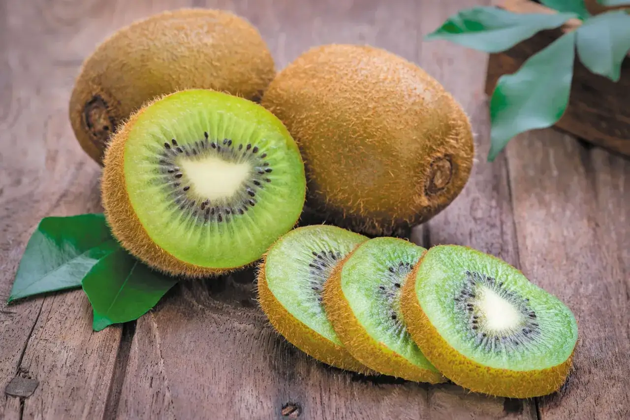 Kiwi jest zdrowe! Na drewnianym blacie leżą całe i pokrojone owoce kiwi, otoczone zielonymi liśćmi.