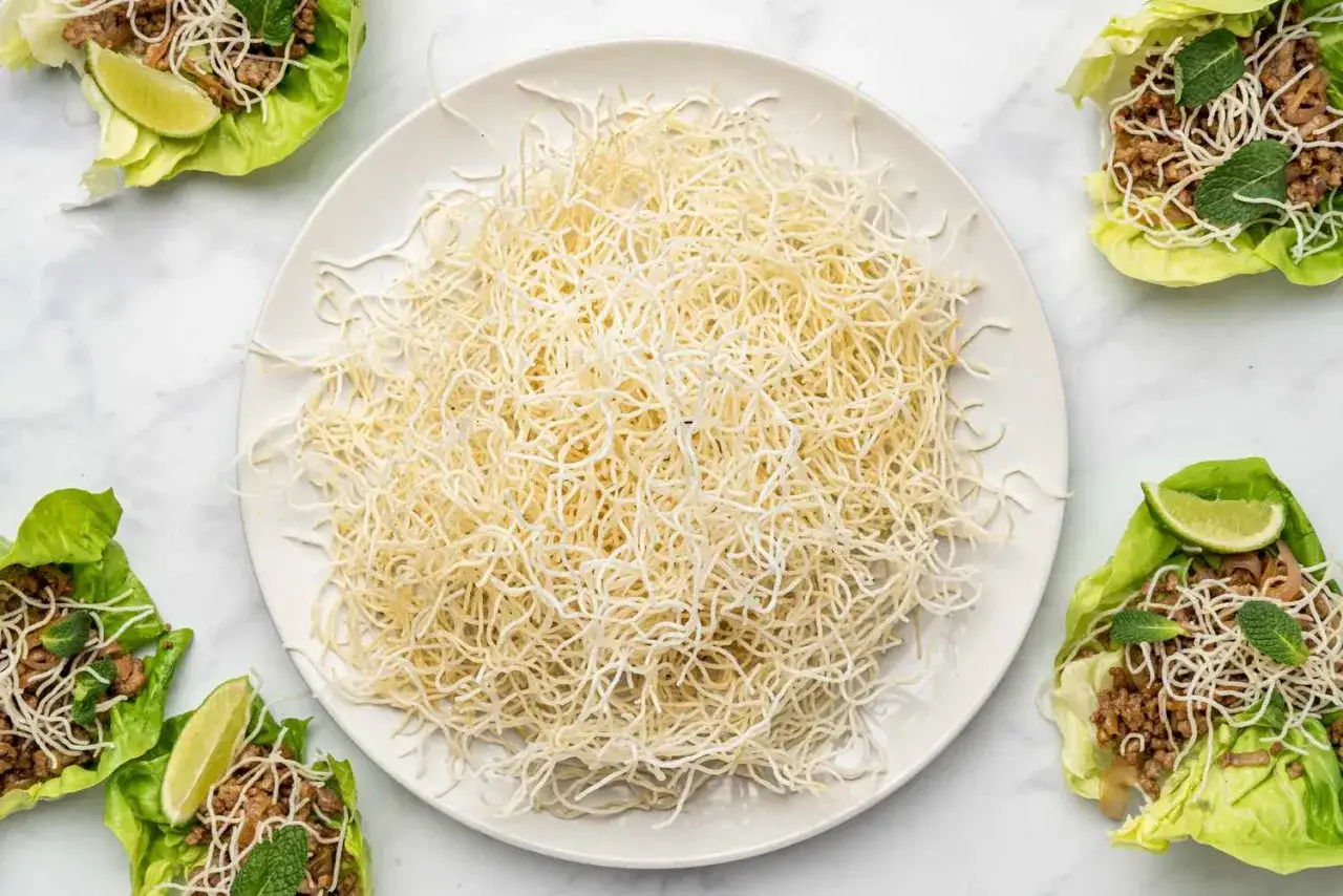 En hög med krispiga, friterade risnudlar på en tallrik, bredvid salladsblad fyllda med köttfärs, mynta och lime.