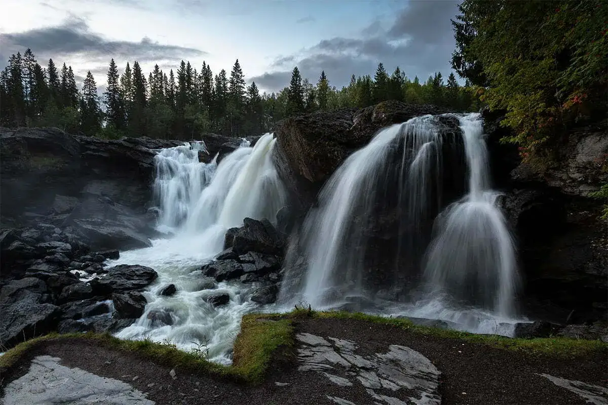 Vattenfall i Norrland, med frodig skog och m&ouml;rka klippor. Vattnet forsar ner i en dramatisk kaskad.