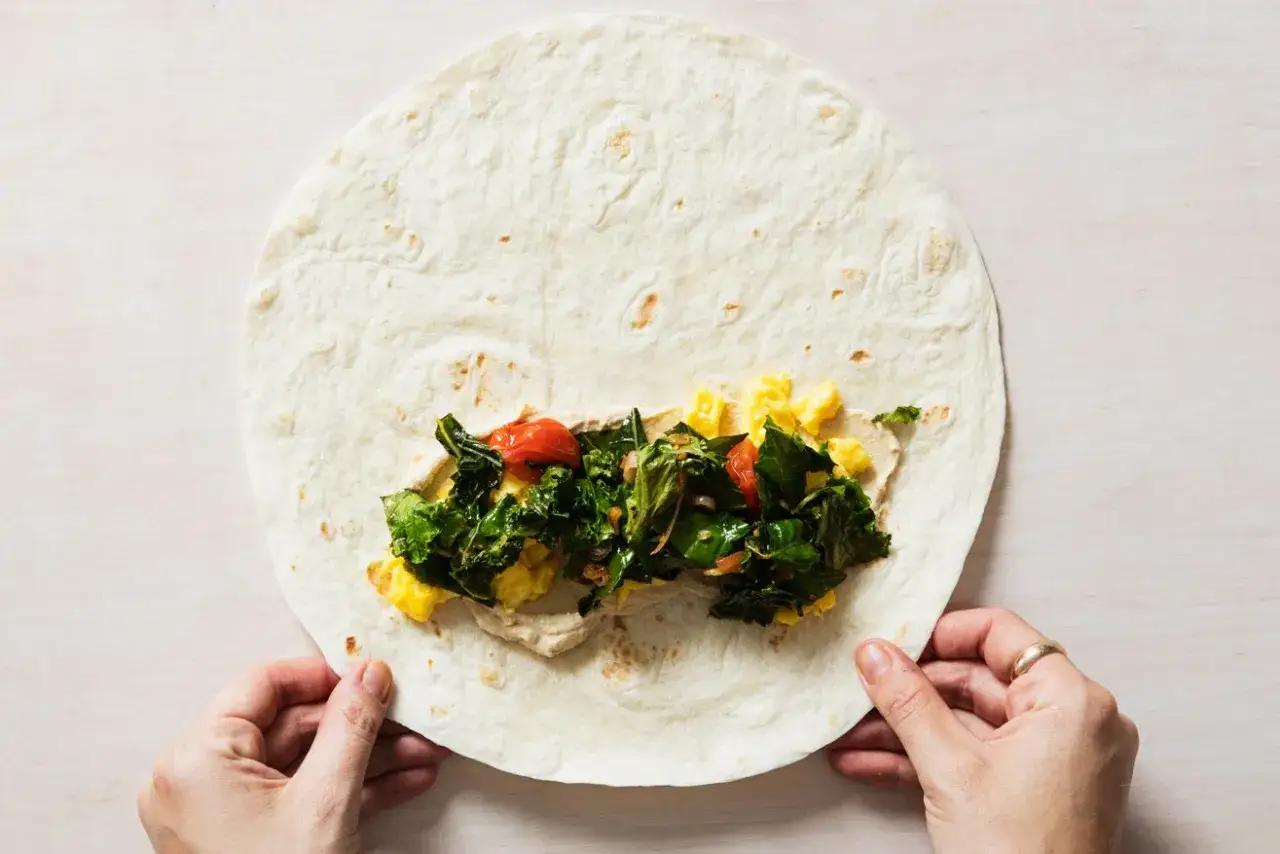 Hände falten einen Wrap mit Rührei, Grünkohl und Tomaten.