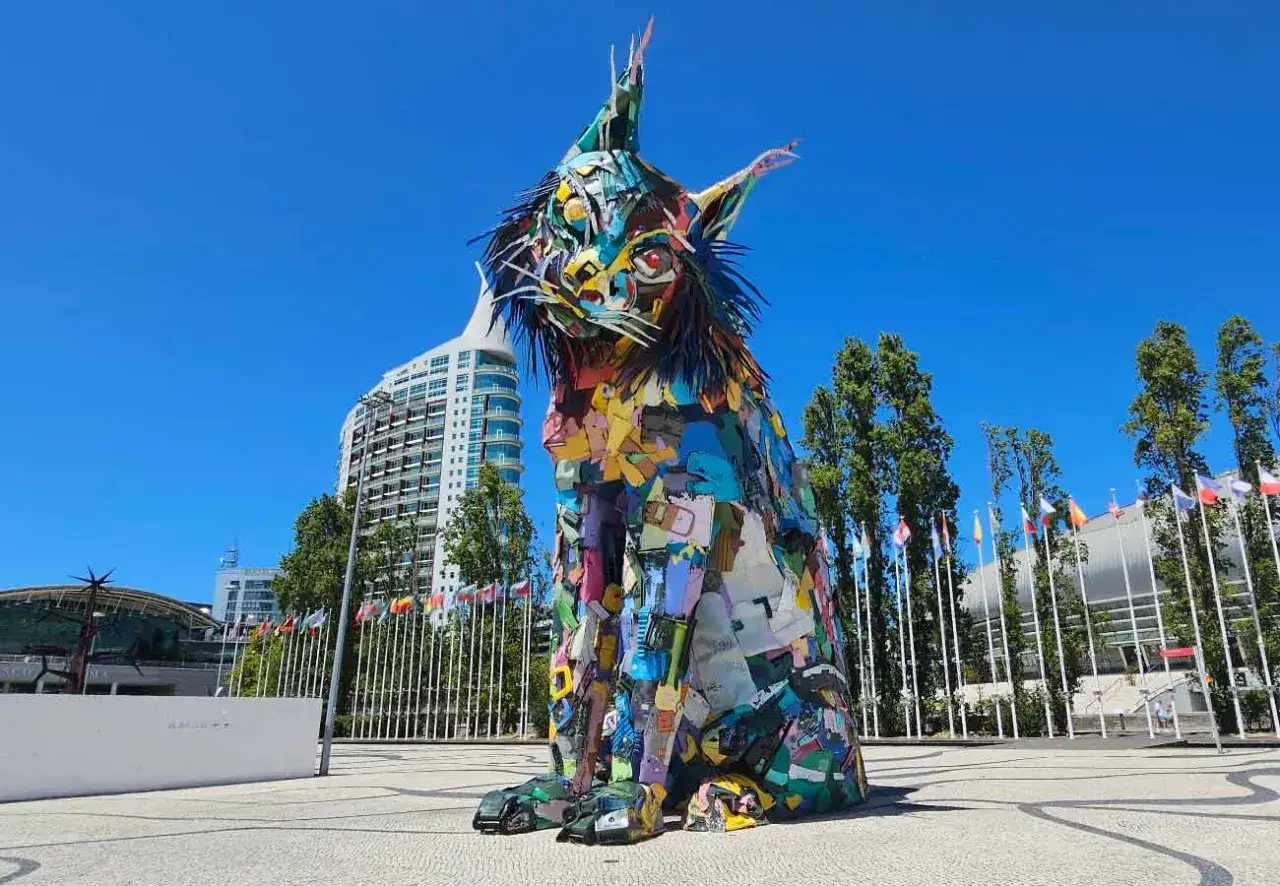 Bunte Luchs-Skulptur aus Recyclingmaterial auf der Expo Lissabon. Hinter ihr wehen Fahnen vor modernen Geb&auml;uden.