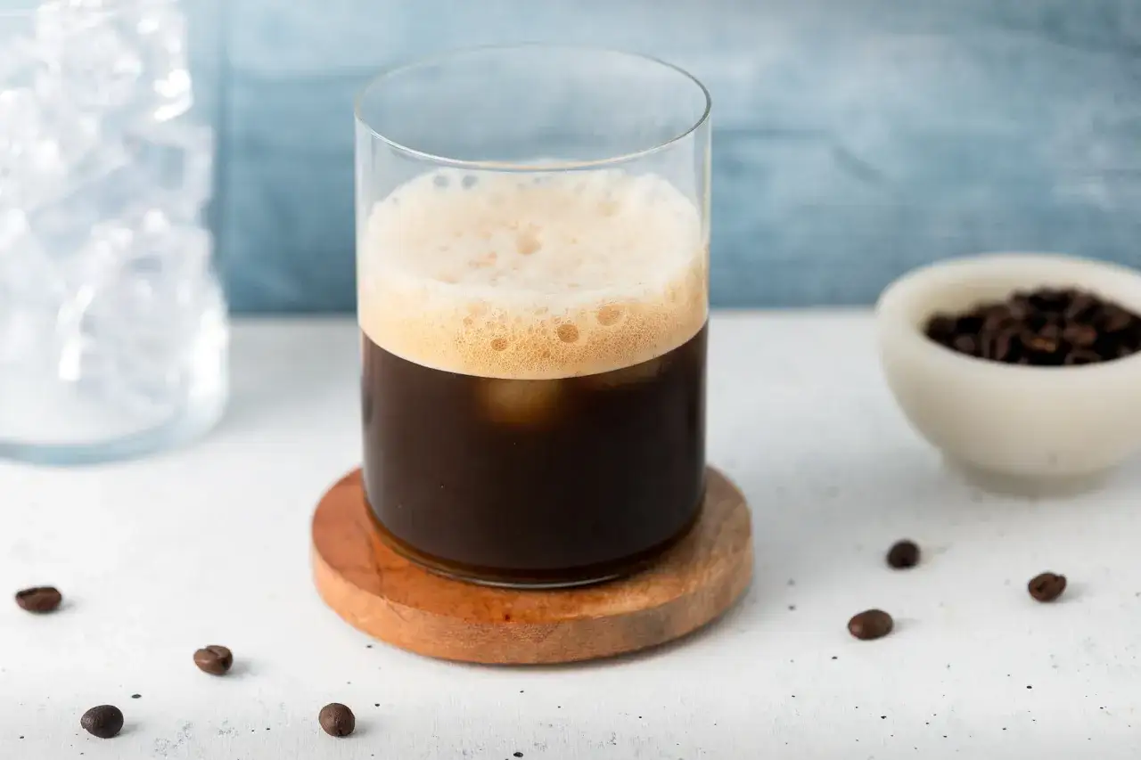 Un bicchiere di caffè shakerato cremoso con schiuma dorata, cubetti di ghiaccio e chicchi di caffè tostati.