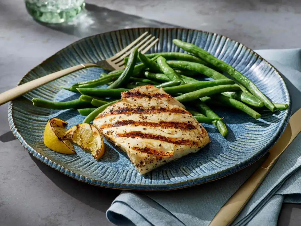 Perfekt gegrillter Fisch mit grünen Bohnen und Zitronenspalten auf einem blauen Teller. Ein köstliches Gericht, das Lust auf Fisch grillen macht.