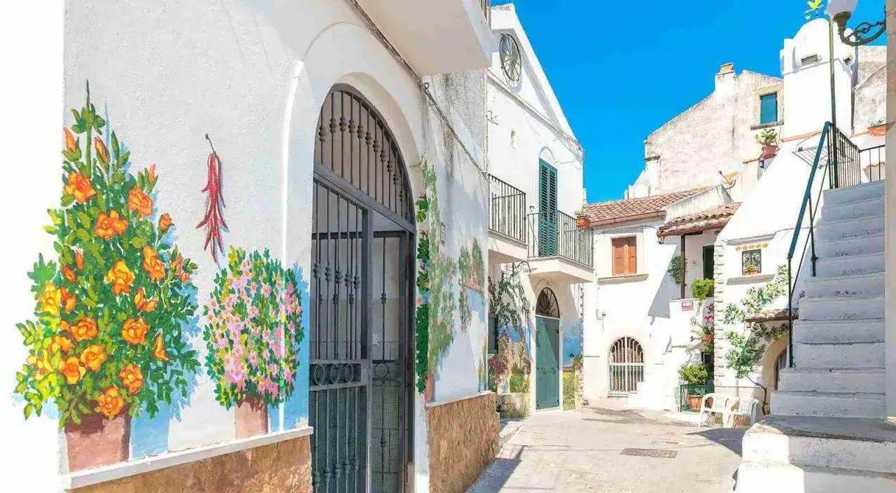 Vicolo fiorito nel centro storico di Rodi Garganico, con murales colorati su muri bianchi e un cielo azzurro brillante.