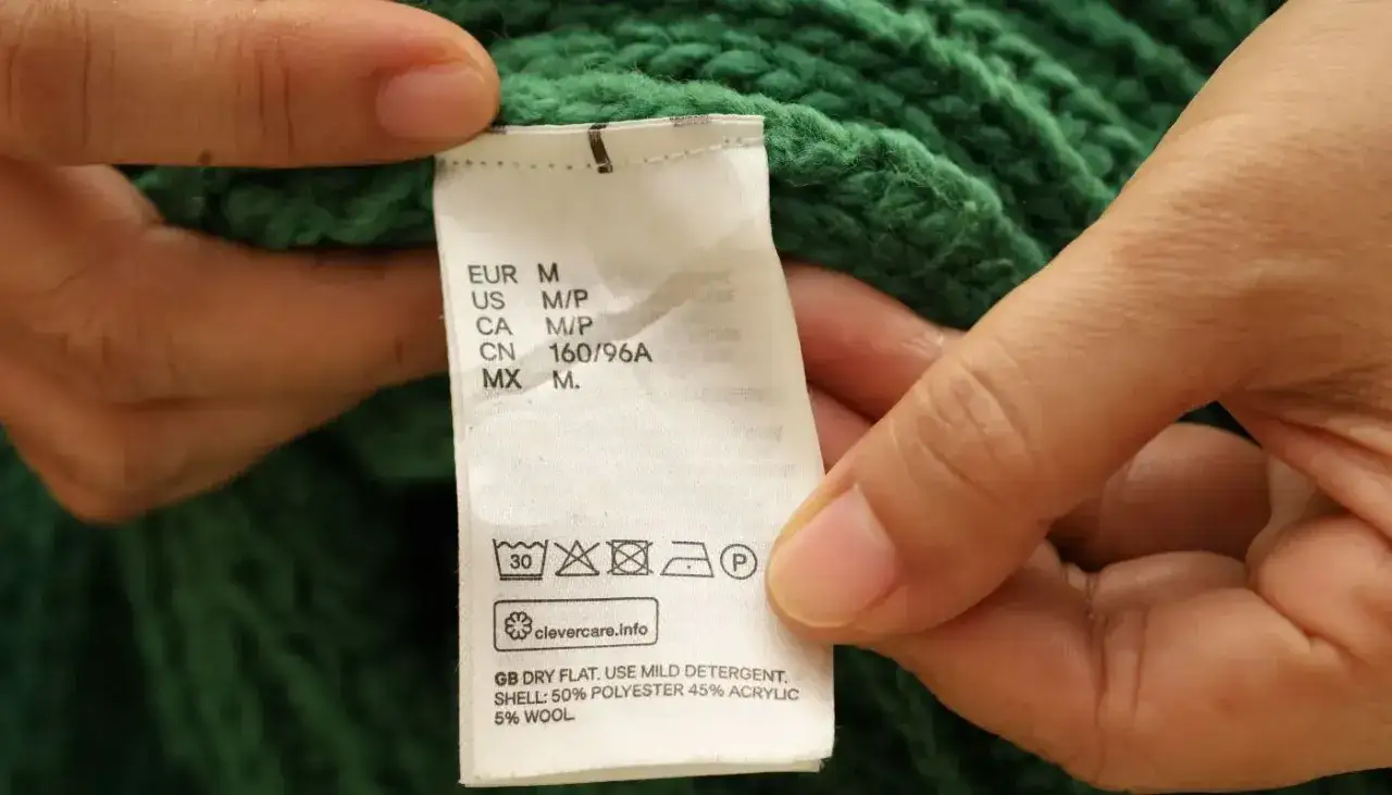 Etichetta di un maglione verde: 50% poliestere, 45% acrilico, 5% lana. Si lava a 30°, non candeggiare, asciugare in piano.