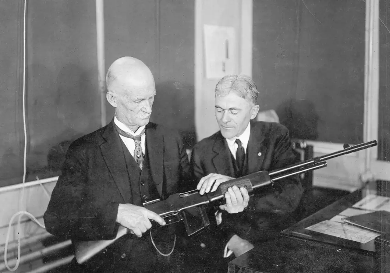 Dw&oacute;ch mężczyzn ogląda karabin. Jeden z nich, z łysą głową, to prawdopodobnie sam John Browning, legendarny konstruktor broni.