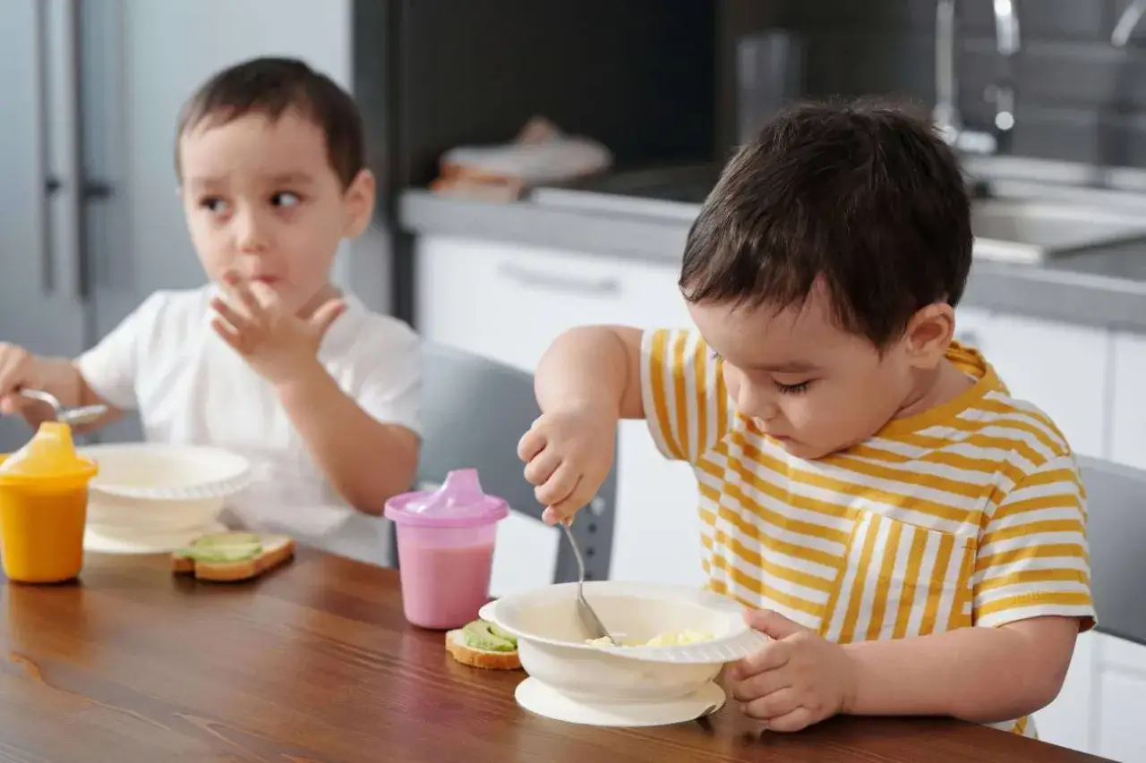 Dos ni&ntilde;os peque&ntilde;os comen en la mesa. El de la derecha, con camiseta a rayas, usa una cuchara para comer de un cuenco. El de la izquierda, con camiseta blanca, se lleva comida a la boca con los dedos. Ideas de recetas para ni&ntilde;os.