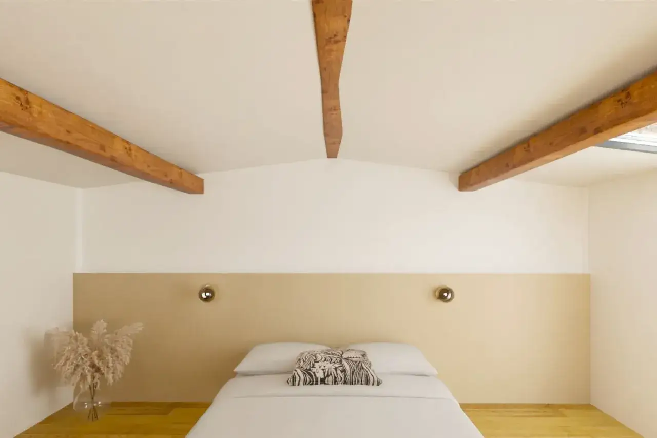 Chambre sous pente avec poutres apparentes. Une t&ecirc;te de lit beige et un lit blanc invitent au repos. Une id&eacute;e peinture pour une chambre sous pente chaleureuse.