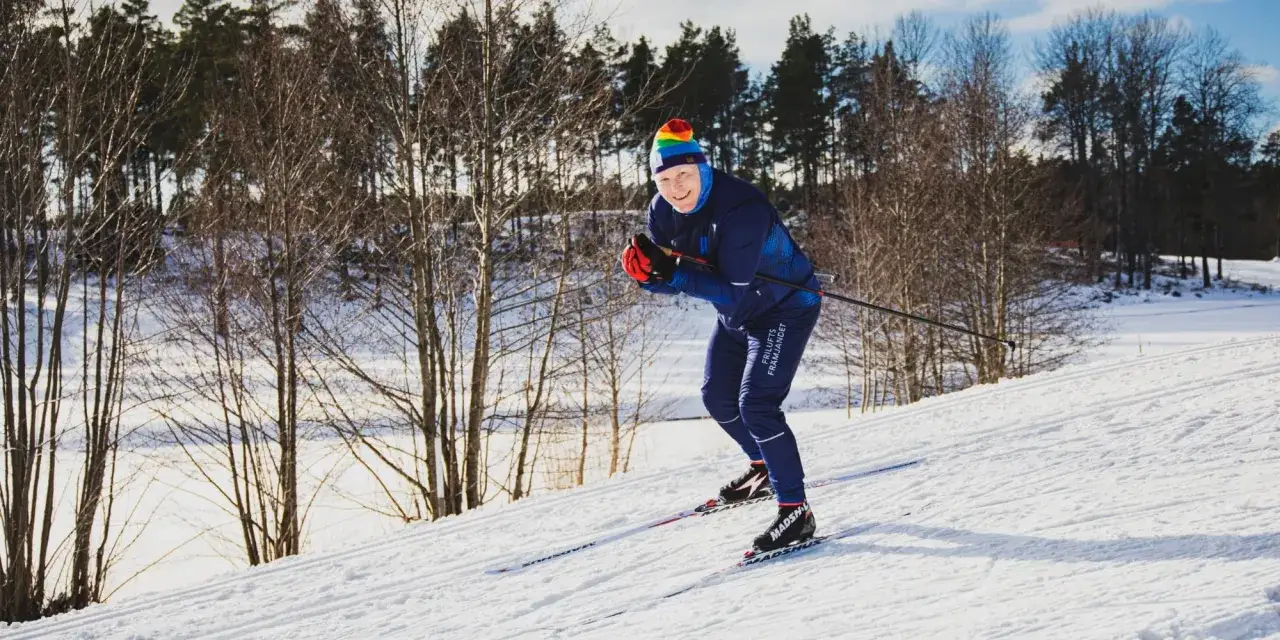 Glad skidåkare i regnbågshatt njuter av ett långlopp skidor i solig vinterterräng.