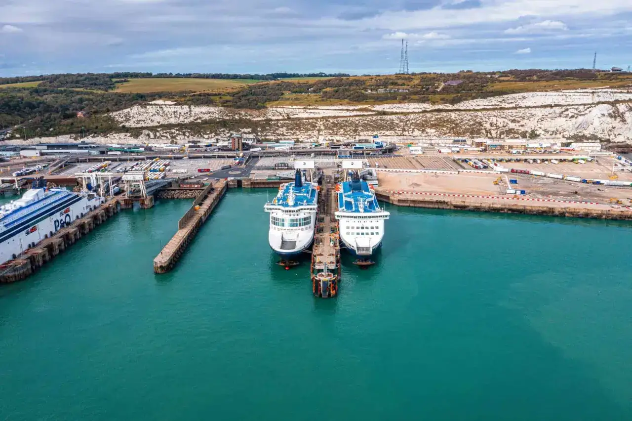 Due traghetti bianchi e blu attraccati nel porto di Dover, con scogliere bianche sullo sfondo e un cielo nuvoloso.