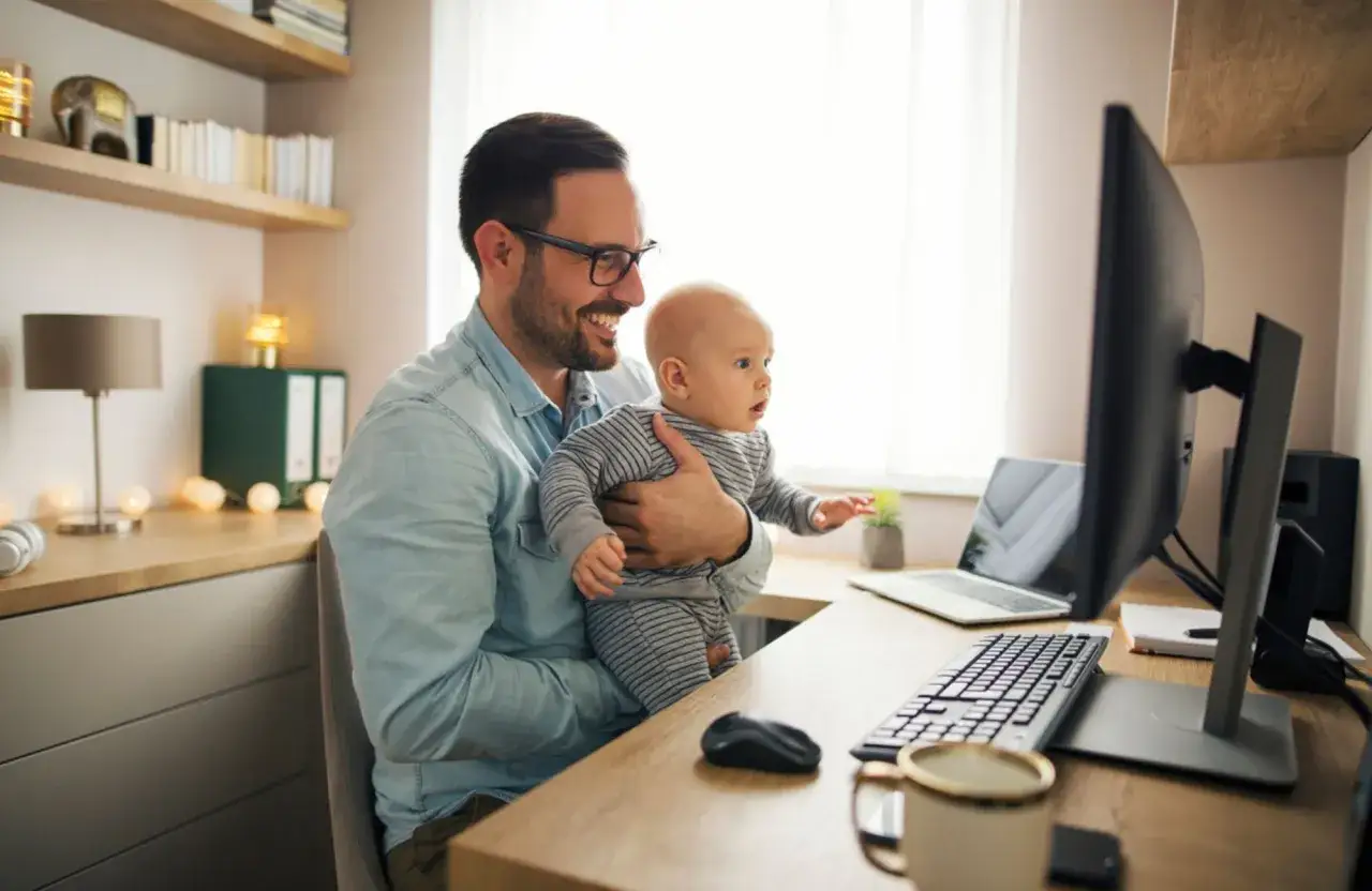Un père souriant tient son bébé tout en travaillant à son bureau. Il semble qu'il prépare sa lettre congé paternité.