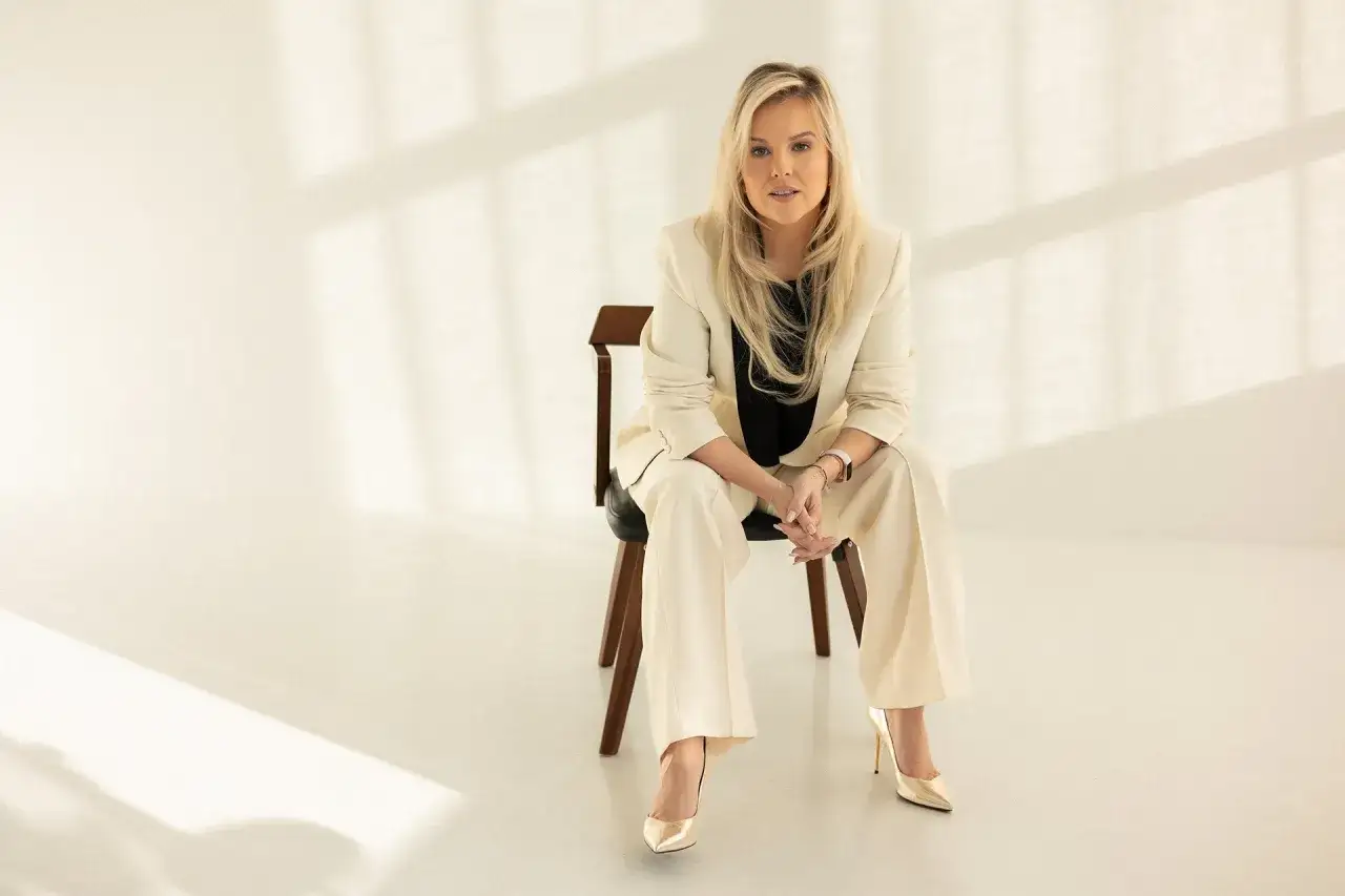 Kamila Kalinczak w eleganckim kremowym garniturze siedzi na krześle, w świetle padającym z okna.