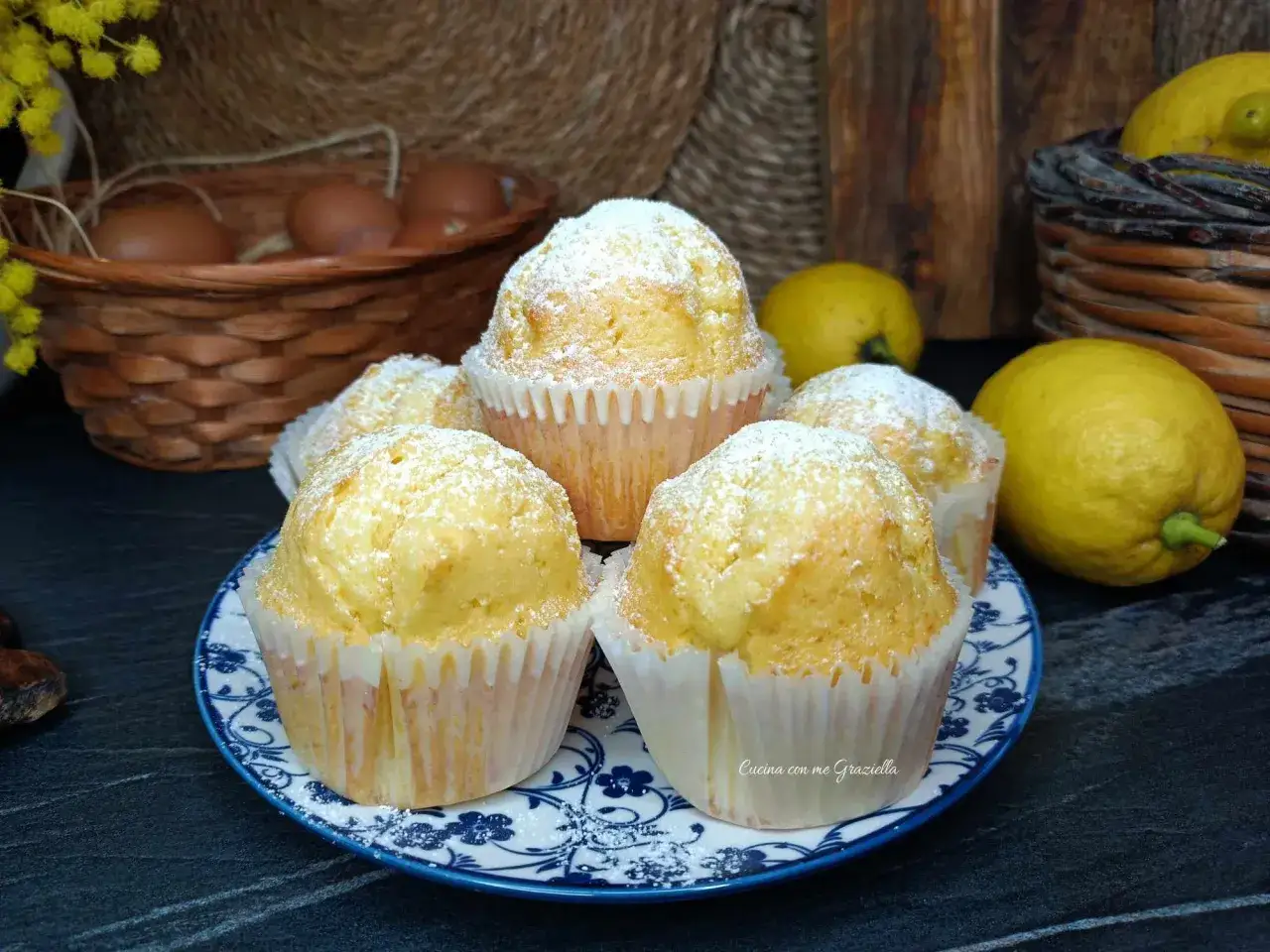 Morbidi muffin allo yogurt, spolverati di zucchero a velo, pronti per essere gustati. Sullo sfondo, uova e limoni freschi.