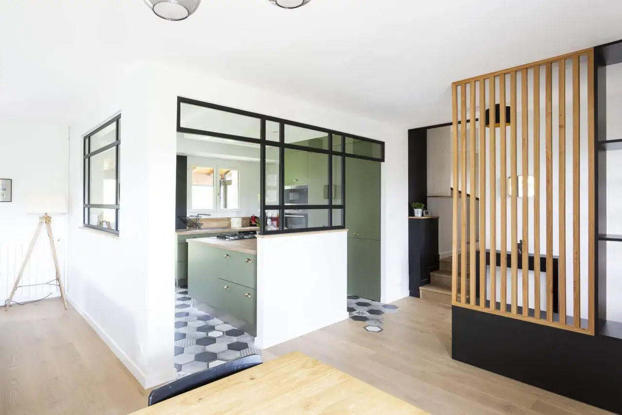 L'entrée de la maison donne directement sur une cuisine moderne aux meubles verts, séparée par une verrière noire. Un escalier en bois est visible.