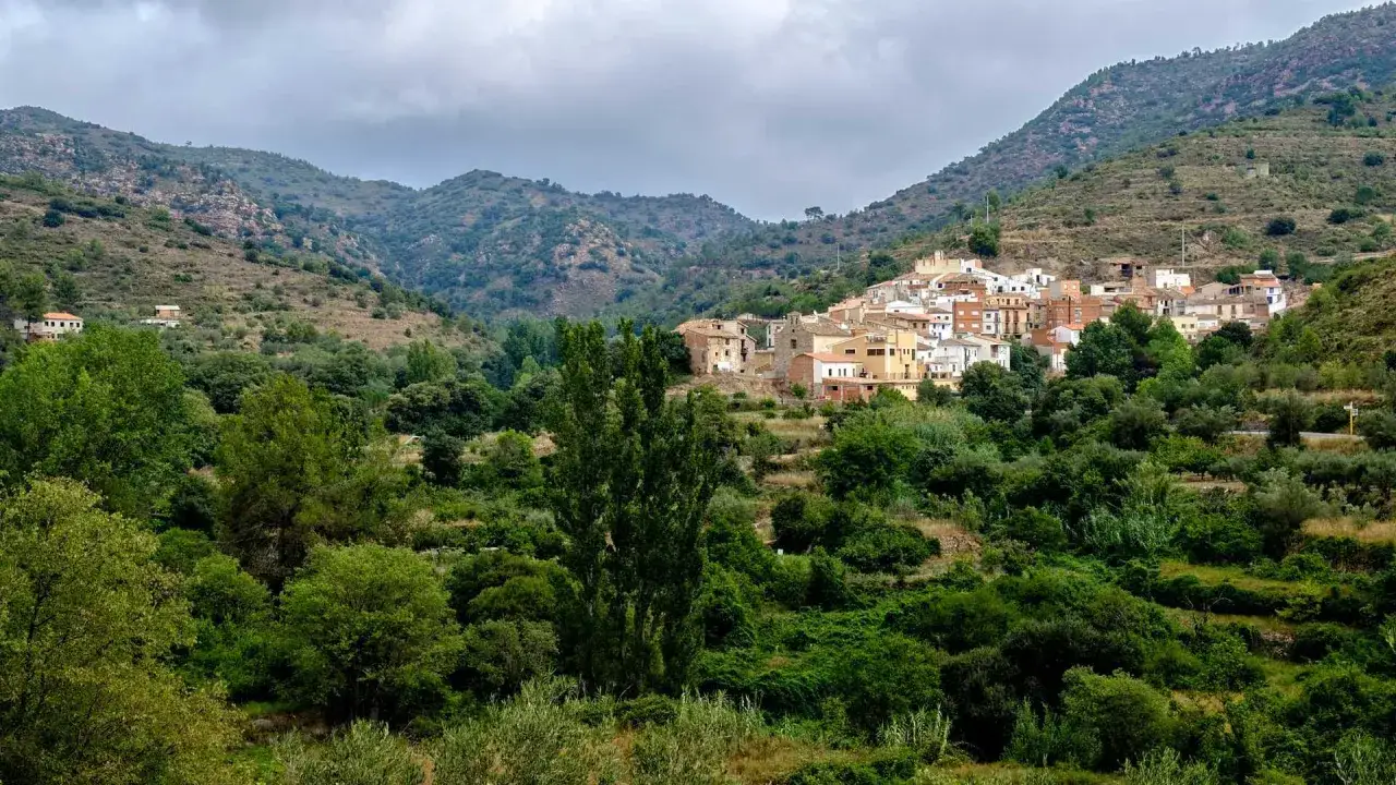 Pueblo blanco anidado en la verde **sierra de Espadán**, rodeado de vegetación exuberante y colinas onduladas bajo un cielo nublado.