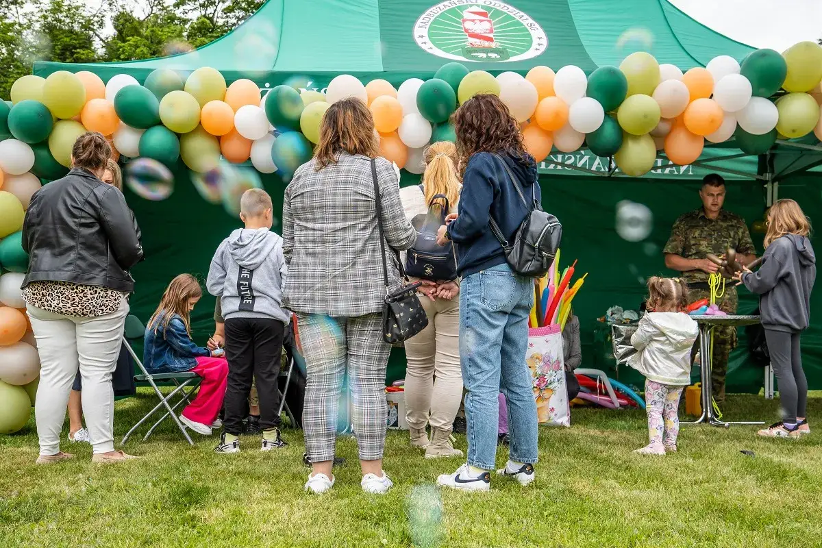 1 czerwca. Dzieci i dorośli pod zielonym namiotem, balony tworzą łuk. Żołnierz tworzy figurki z balonów dla dzieci.