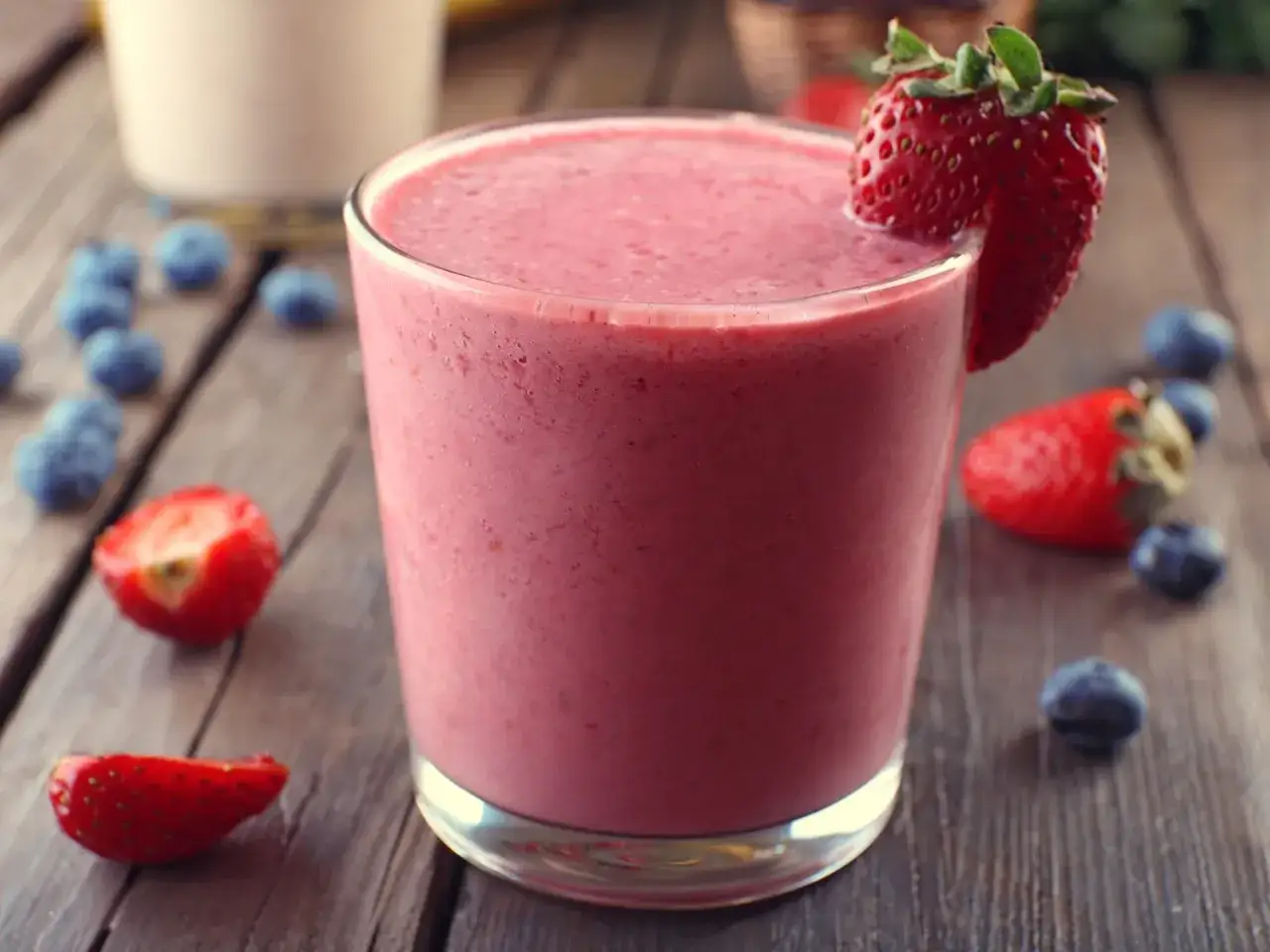 Un d&eacute;licieux smoothie fraise banane, garni d'une fraise fra&icirc;che, entour&eacute; de fruits rouges sur une table en bois.
