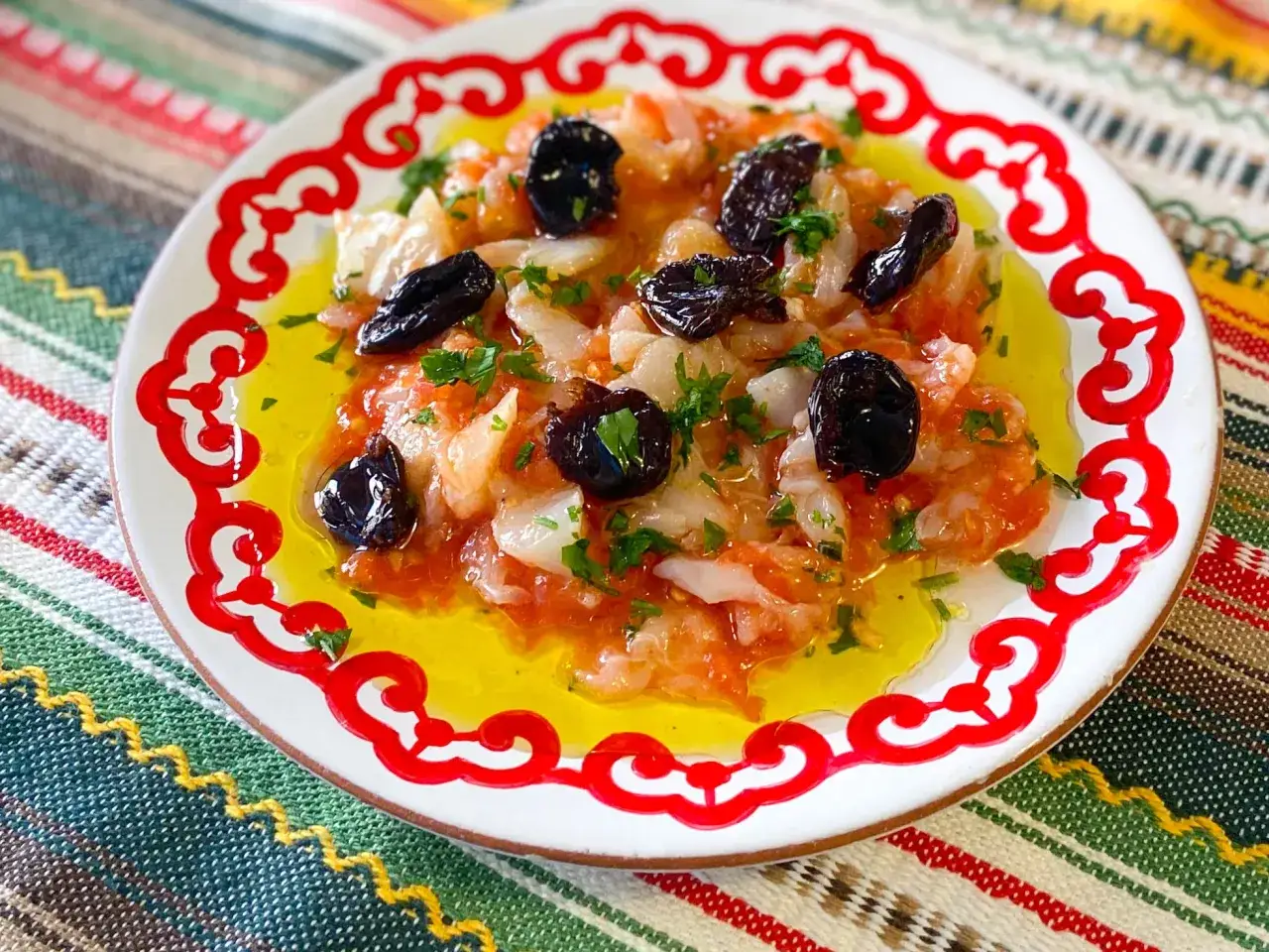Deliciosa ensalada de bacalao con aceitunas negras y perejil, bañada en aceite de oliva, servida en un plato blanco con borde rojo.