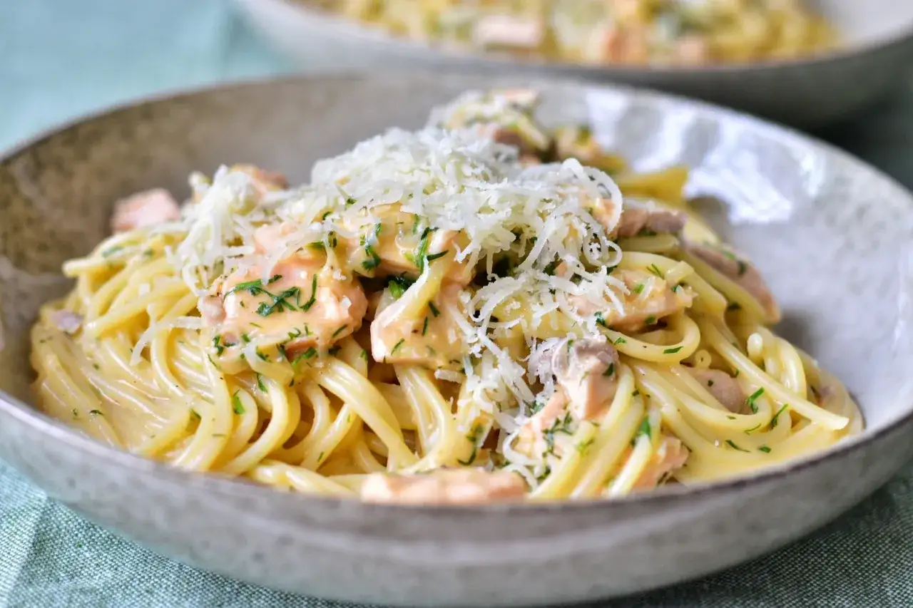 En n&auml;rbild av en sk&aring;l med kr&auml;mig laxpasta, toppad med riven ost och f&auml;rsk persilja. Ett perfekt laxpasta recept f&ouml;r en snabb middag.