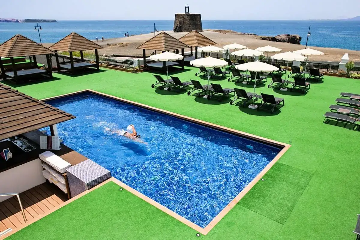 Disfruta de viajes a Lanzarote todo incluido 2x1. Una persona nada en una piscina azul, rodeada de césped artificial, tumbonas y el mar al fondo.