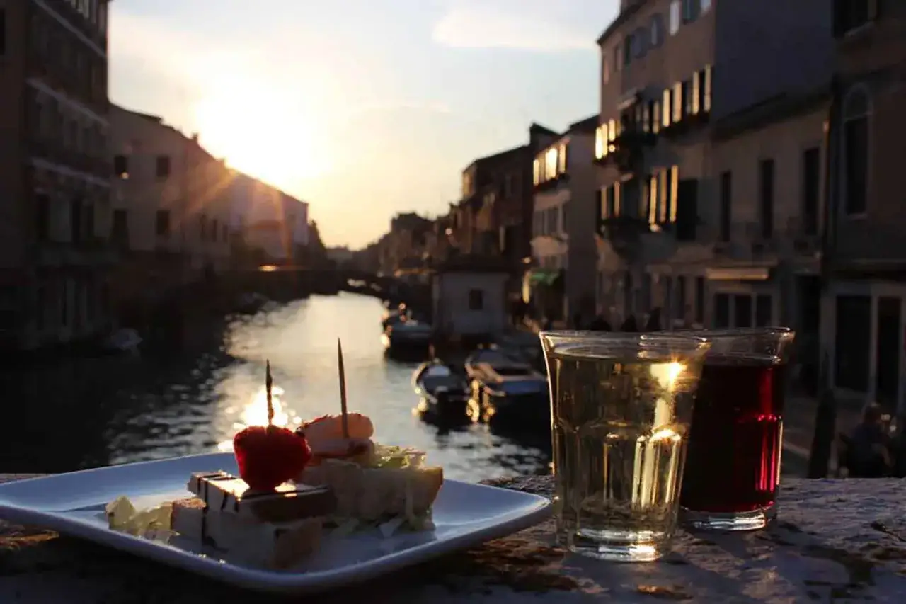 Un aperitivo al tramonto a Venezia, con due bicchieri di vino e stuzzichini su un canale, un momento perfetto per innamorati a Venezia.