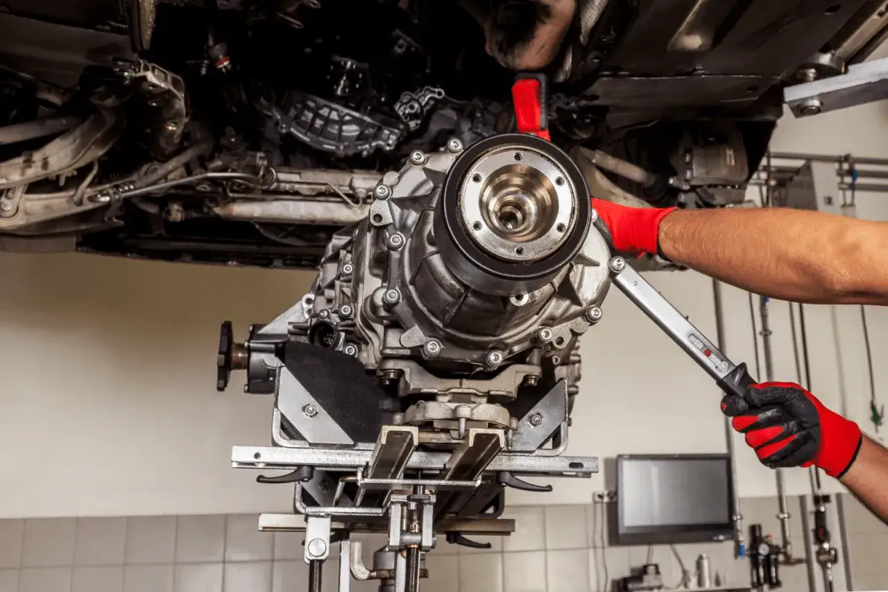 Mechanik w czerwonych rękawicach dokonuje wymiany skrzyni bieg&oacute;w w samochodzie na podnośniku.