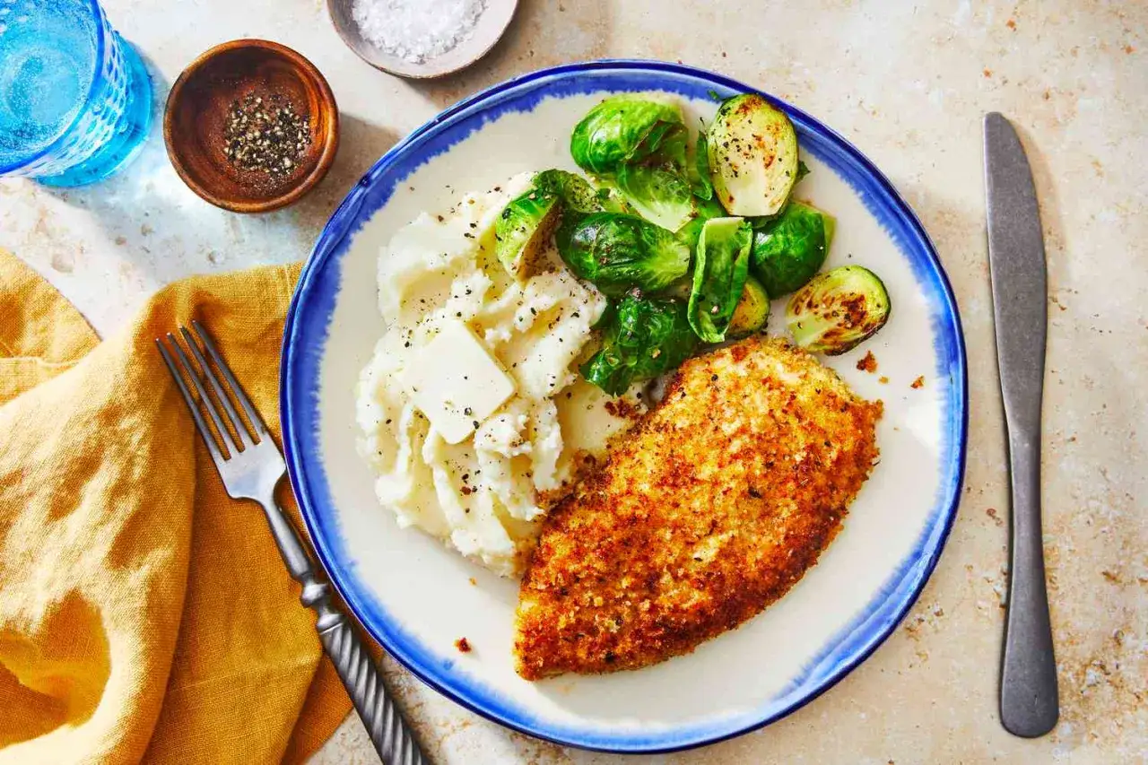 Une escalope de poulet panée dorée, servie avec une purée de pommes de terre crémeuse et des choux de Bruxelles rôtis.