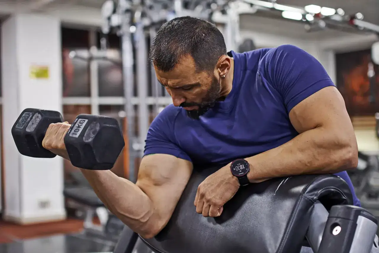 Man med skägg utför preacher curls med hantlar på ett gym.
