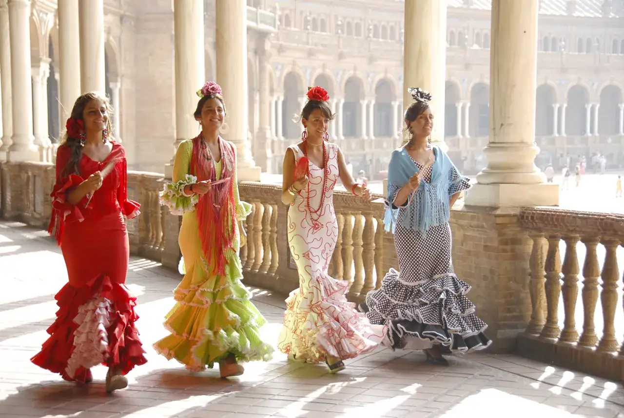 Cuatro sevillanas, vestidas con trajes de volantes de colores vibrantes, pasean sonrientes.