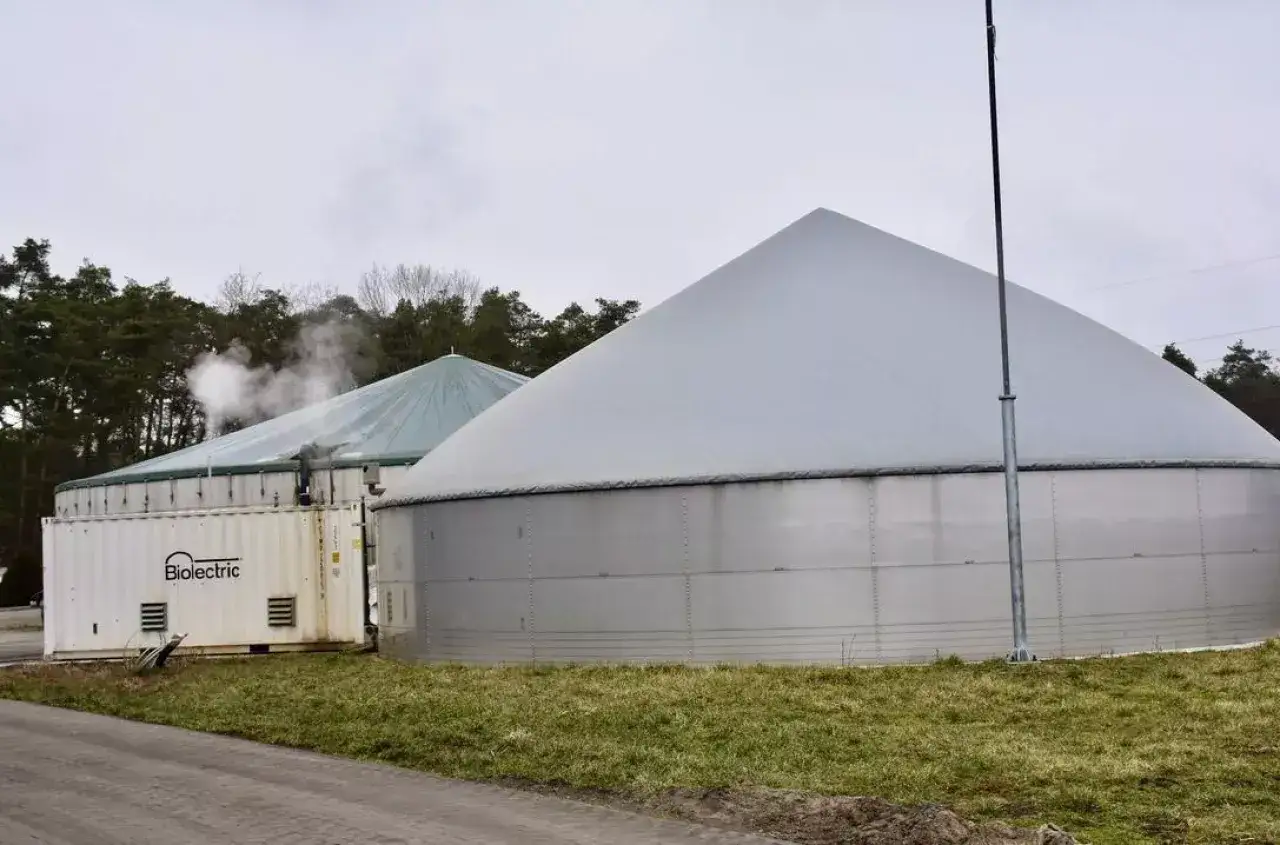 Dwa wielkie zbiorniki biogazowni, przetwarzającej surowce energetyczne. W tle las, na pierwszym planie trawa.