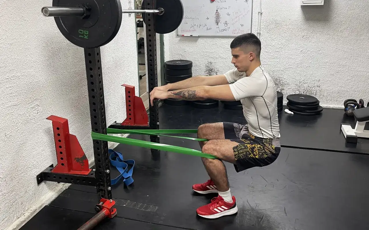 Mężczyzna wykonuje ćwiczenie spanish squat z gumą oporową wokół kolan, w siłowni.