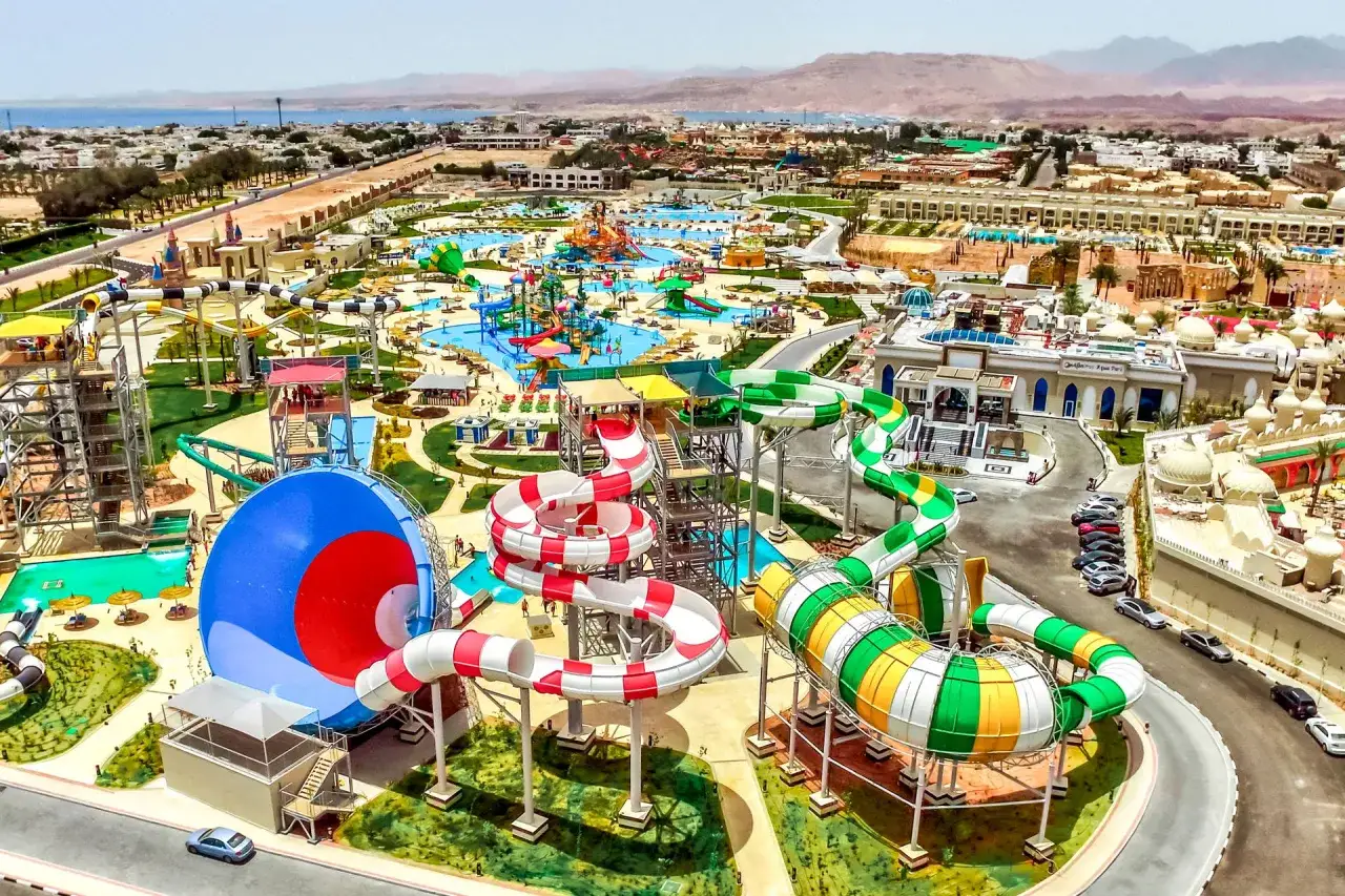 Kolorowe zjeżdżalnie wodne i baseny w Albatros Aqua Park, otoczone budynkami hotelowymi i górami w tle.