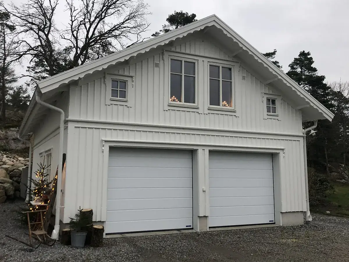 Vitt garage med snedtak och två portar. En julgran med ljus står vid sidan.