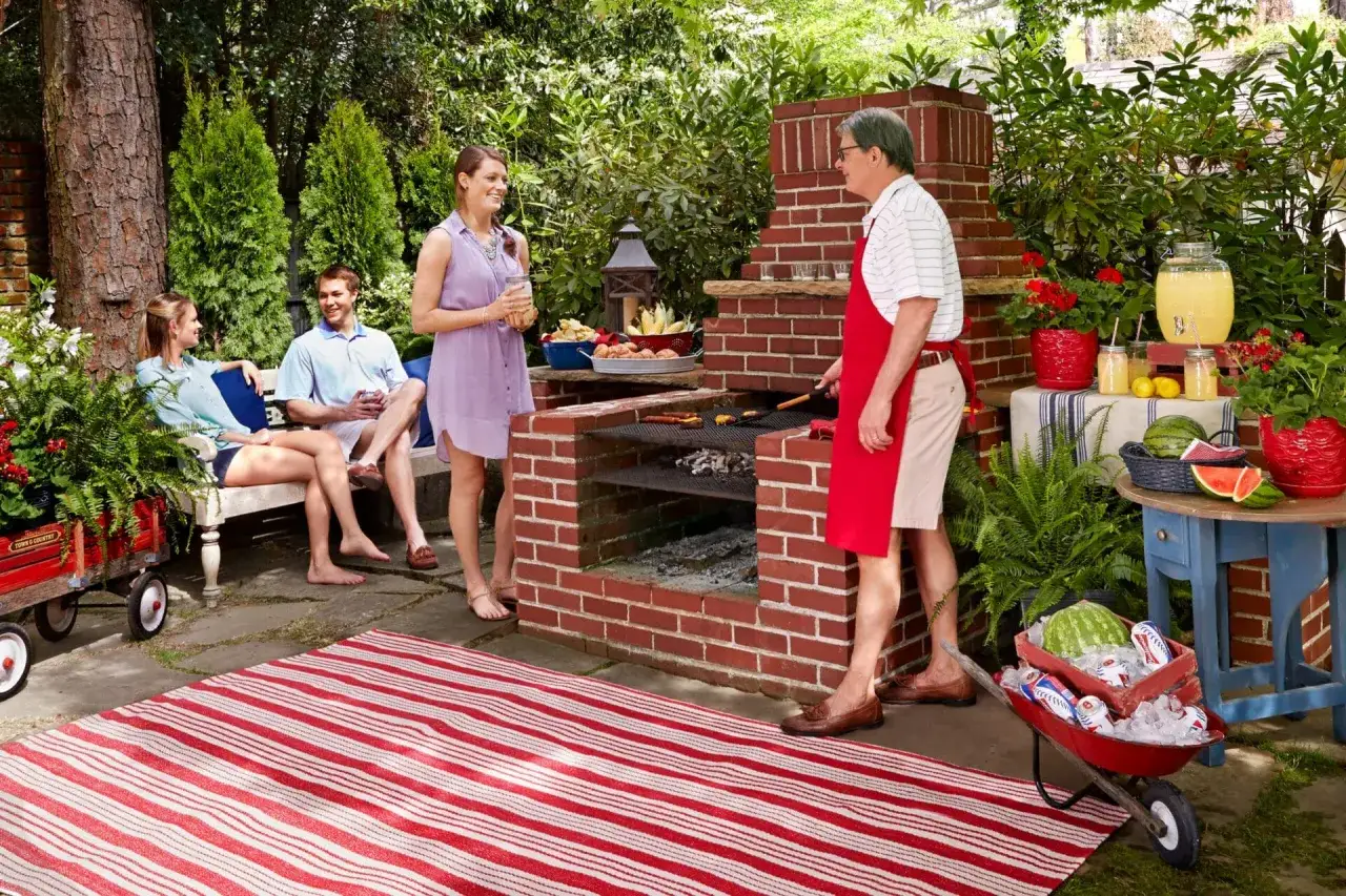 Familj njuter av en grillfest vid en murad grill. En man grillar, medan andra sitter och pratar.