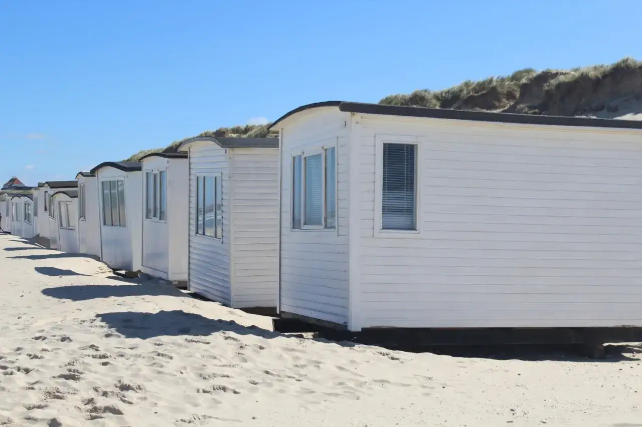 Rząd białych domków holenderskich na piaszczystej plaży, z wydmami w tle i błękitnym niebem.