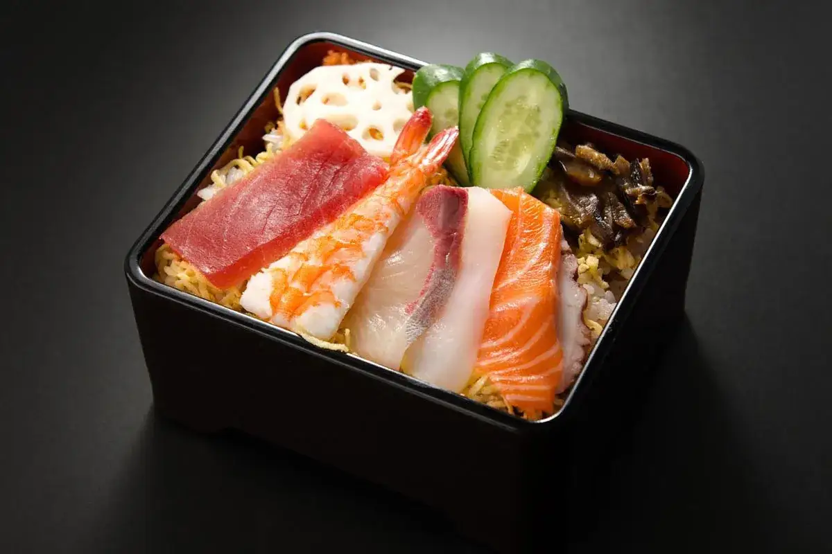 Chirashi zushi: kolorowe plastry ryb (łosoś, tuńczyk, biała ryba), krewetka, plasterki ogórka i kwiat lotosu na ryżu.