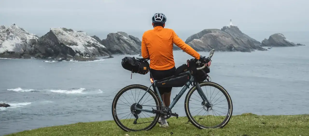 Rowerzysta w pomarańczowej kurtce z sakwami na wyprawę bikepacking gravel podziwia surowe wybrzeże z latarnią w oddali.